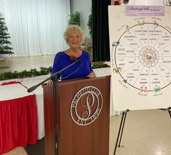 A woman with curly hair stands at a podium, smiling, next to a chart displaying astrological signs.